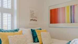 Een slaapkamer met een bed, kussens, een kunstwerk aan de muur en een raam met zonwering.