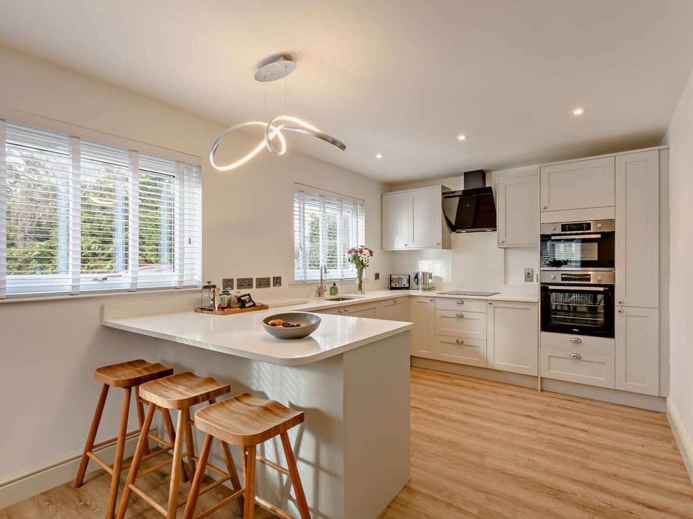 A modern kitchen with a central island, wooden stools, white cabinets, and built-in appliances.
