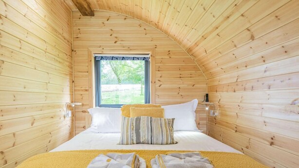 Une pièce chaleureuse aux murs en bois, avec un lit recouvert d’oreillers, une fenêtre et une table de chevet.