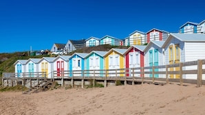 Une rangée de cabanes de plage colorées, séparées par une clôture en bois, se trouve sur une plage de sable.
