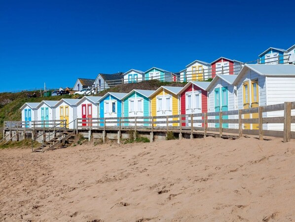 A row of colorful beach huts with a wooden fence in front, situated on a sandy beach. - 1 Bed in Holsworthy (oc-89791) (Holsworthy Hamlets)