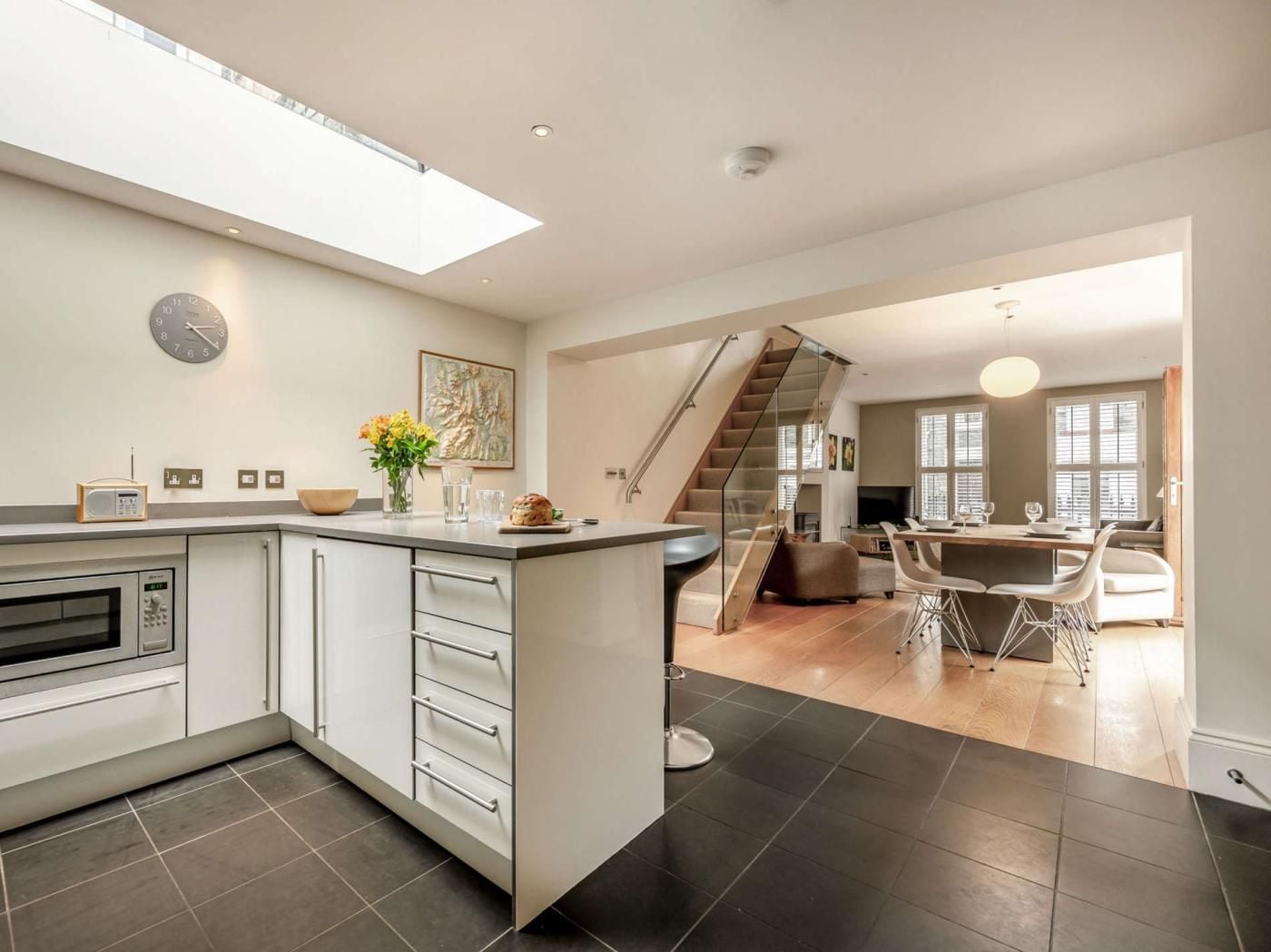 A modern kitchen with a central island, a dining area with a glass table, and a staircase leading to an upper level.