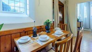Uma sala de jantar com uma mesa de madeira, pratos brancos, uma cafeteira preta e um vaso com planta.