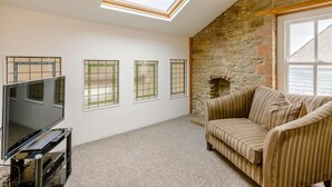 A room with a TV on a stand, a sofa, a stone wall, and three windows with a grid pattern. - 3 Bed in Coleford (oc-89806) (Coleford)