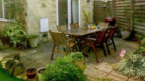 A patio with a wooden table and chairs, surrounded by potted plants and a brick wall. - 3 Bed in Cheltenham (oc-89850) (Bourton-on-the-Water)