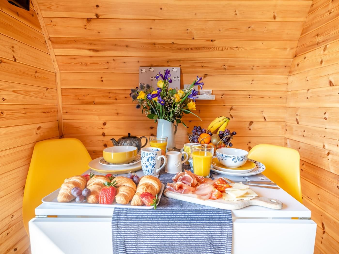 Ein Holzblockhaus-Interieur mit einem Esstisch, der für ein Frühstück gedeckt ist, inklusive Croissants, Obst und verschiedenen Getränken.