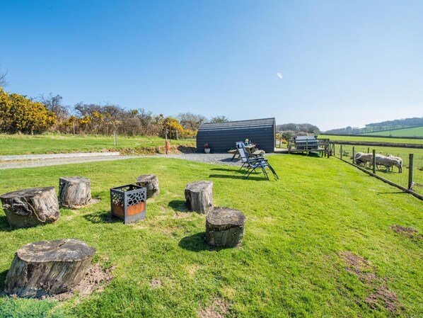 A grassy outdoor area with a wooden structure, a picnic table, and a fenced area with sheep.