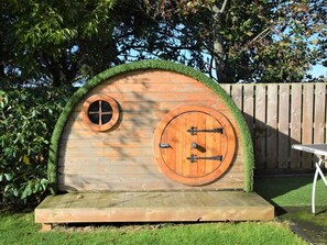 A wooden structure with a circular door and a smaller window, surrounded by a grassy area and a wooden fence. - 1 Bed in Looe (oc-90170) (Trenewan)