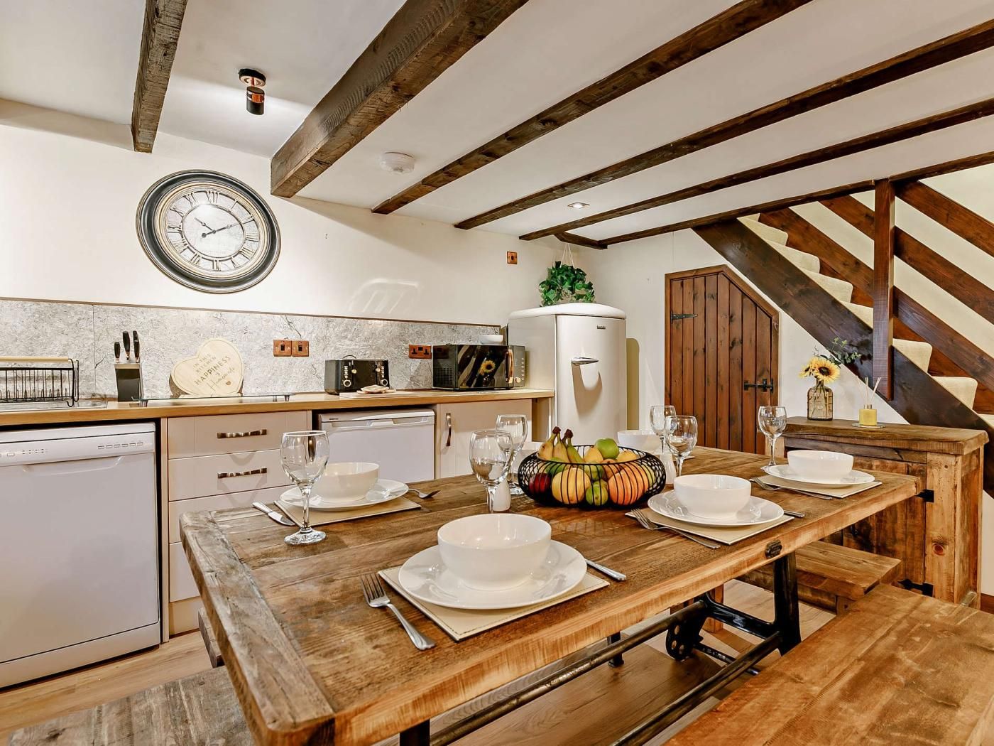 Una cocina rústica con vigas de madera, un reloj antiguo y una mesa de comedor preparada para cuatro.