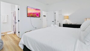 A bedroom with a bed, a TV mounted on the wall, a bedside lamp, and a door leading to another room.