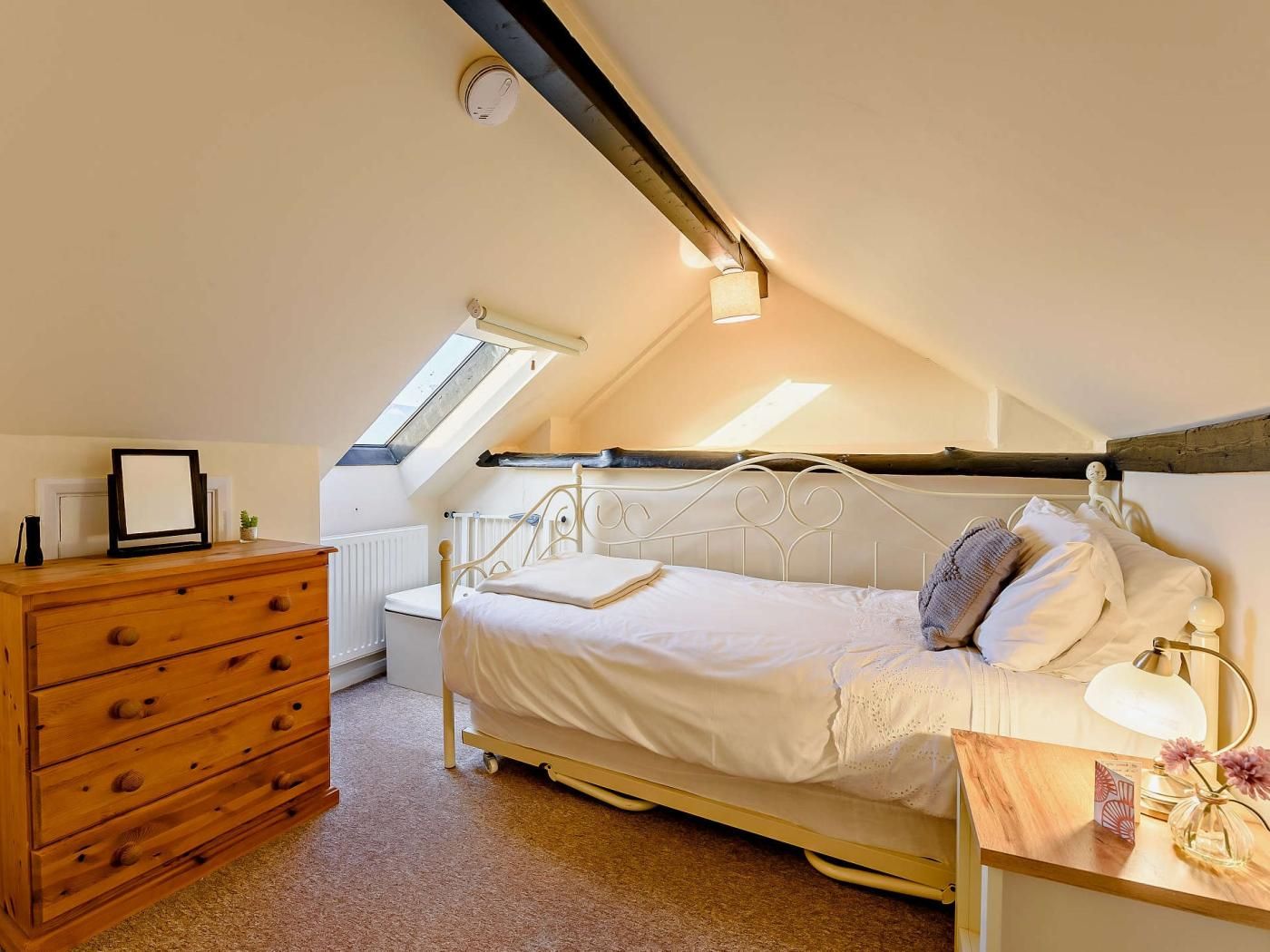 Ein Schlafzimmer mit schrägem Dach, einem Bett mit weißer Bettwäsche, einer Holzkommode, einem Nachttisch mit Lampe und einem kleinen Blumentopf.