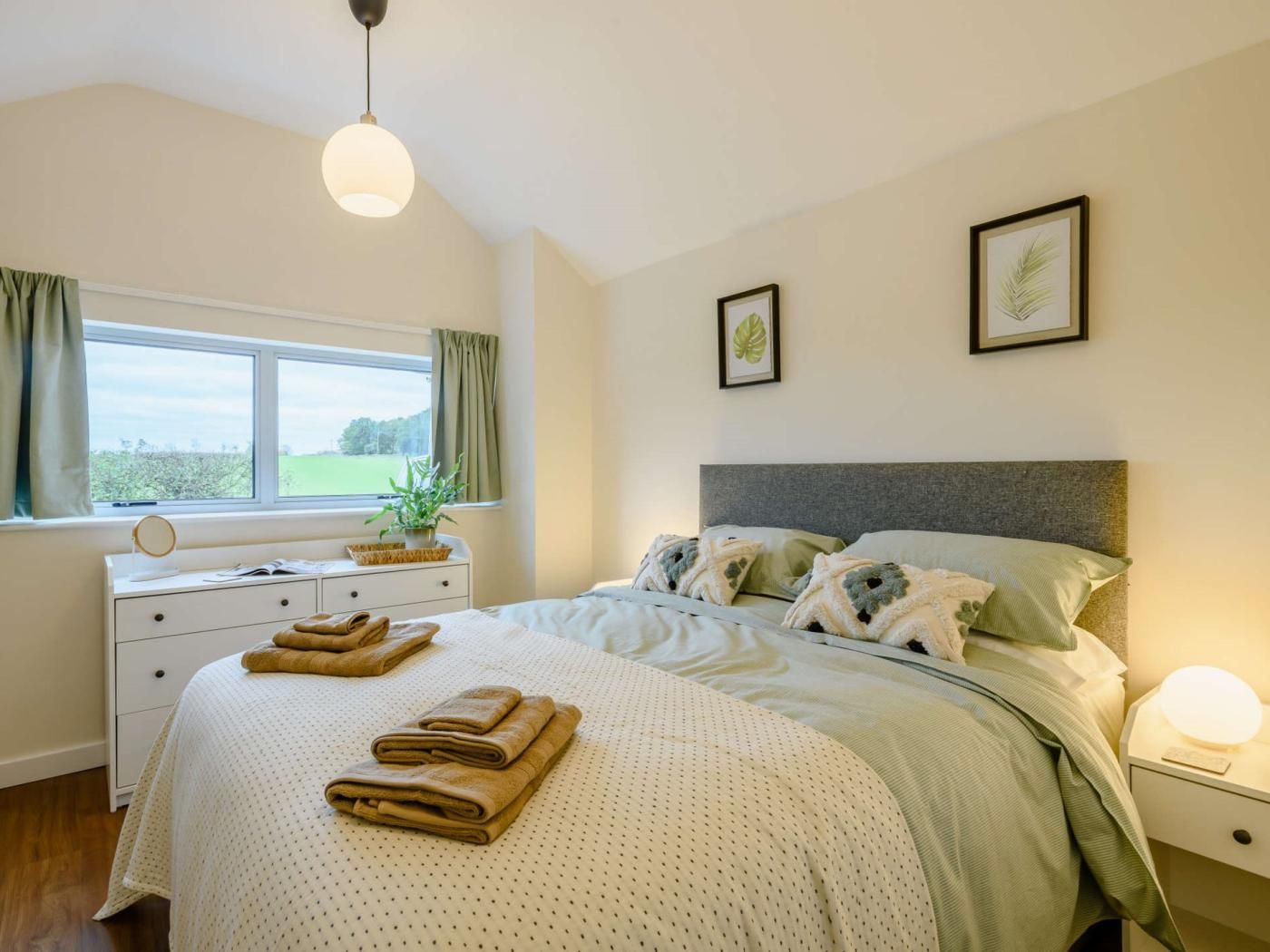 A bedroom with a bed, bedside tables, a window with curtains, a plant, and framed pictures on the wall.