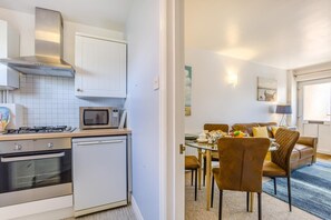 A compact kitchen with a microwave, stove, and dishwasher. Adjacent is a dining area with a table, chairs, and a sofa. - 1 Bed in Aldeburgh (oc-89682) (Aldeburgh)