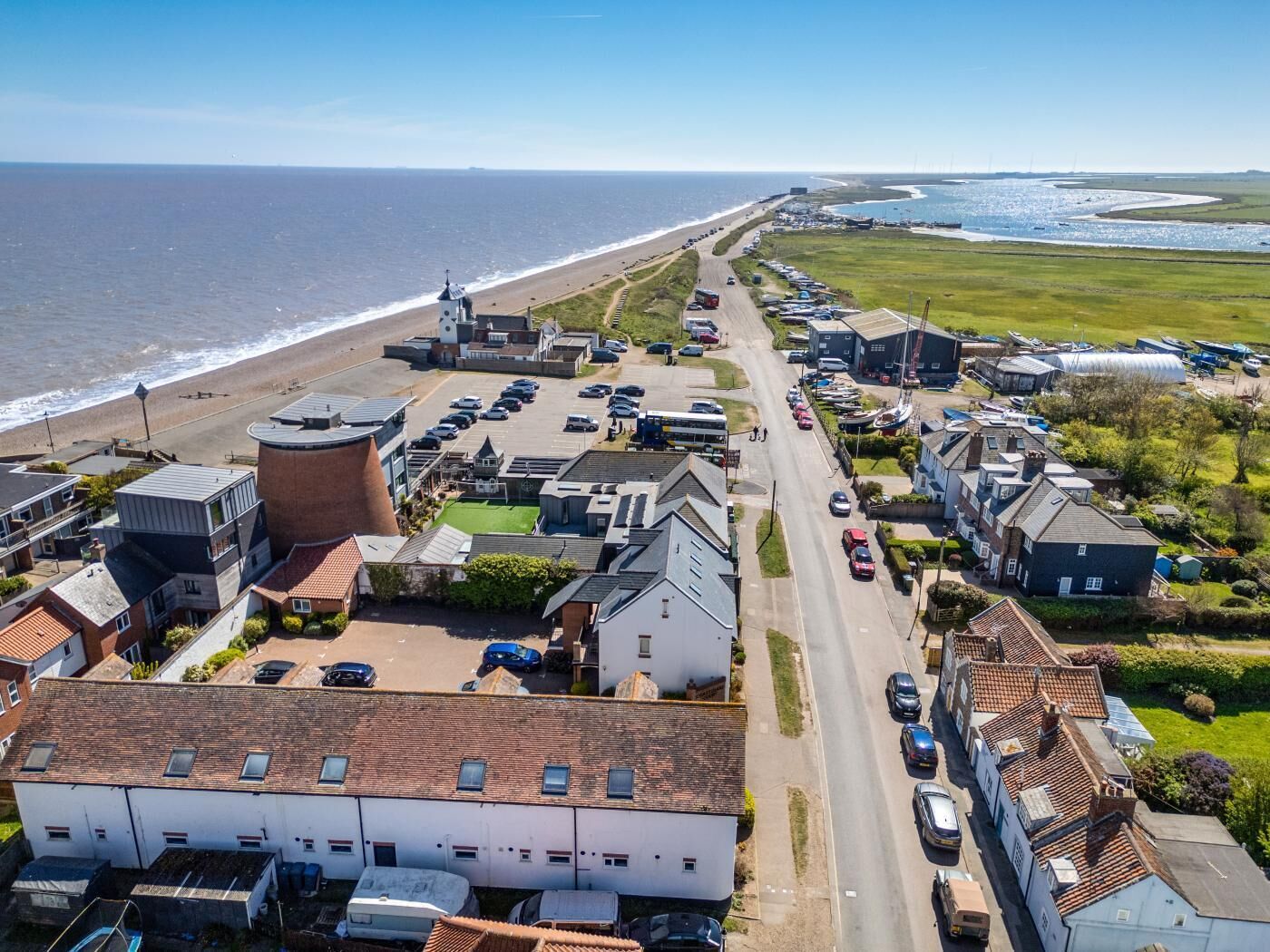 Vue aérienne d’une ville côtière avec une plage, une route bordée de voitures garées et divers bâtiments, notamment des maisons d’habitation et des structures industrielles.