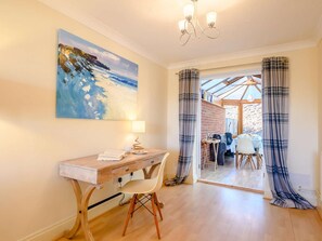 A room with a wooden desk, a chair, a painting on the wall, and a view through a glass door to an outdoor dining area. - 3 Bed in King's Lynn (oc-89756) (Syderstone)