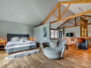 Quarto moderno com uma cama grande, uma poltrona cinza, vigas de madeira, piso de madeira e vista para a sala de estar.