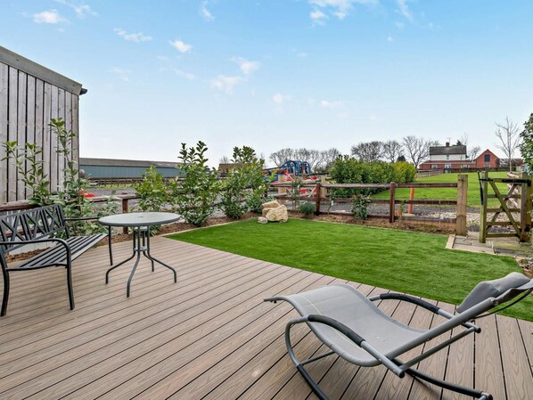 A wooden deck with a table and chairs, a grassy lawn, and a playground in the background.