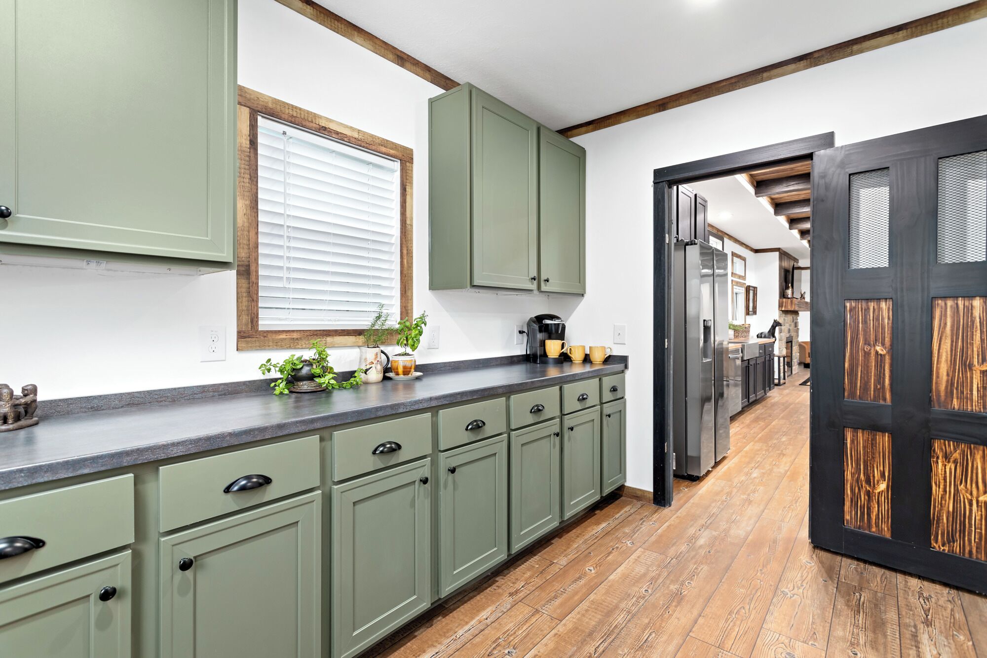 Une cuisine avec des meubles de rangement verts, une fenêtre avec des stores, une machine à café et une porte avec un cadre en bois.