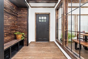 Un pasillo con piso de madera, una puerta oscura, un banco y una mesa con sillas.