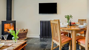 Et hyggeligt rum med brændeovn, spisebord med stole, en radiator og et fladskærms-tv monteret på væggen.