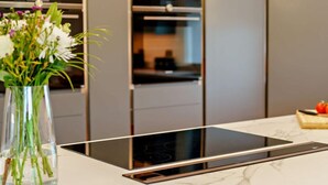 Una cocina moderna con una placa de inducción integrada, un florero transparente con flores y una tabla de cortar de madera con tomates.