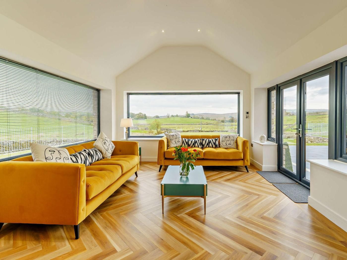 A spacious living room with two yellow sofas, a coffee table, and large windows offering a view of the countryside.