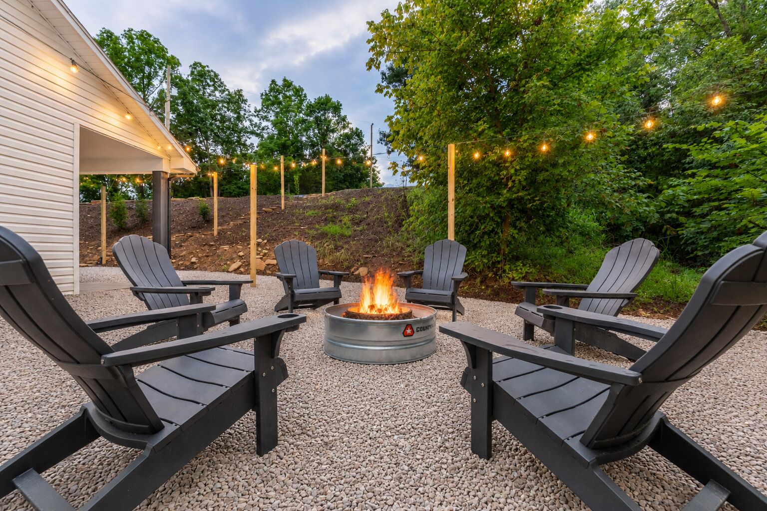 Un foyer entouré de fauteuils Adirondack, avec un sol en gravier et des guirlandes lumineuses.