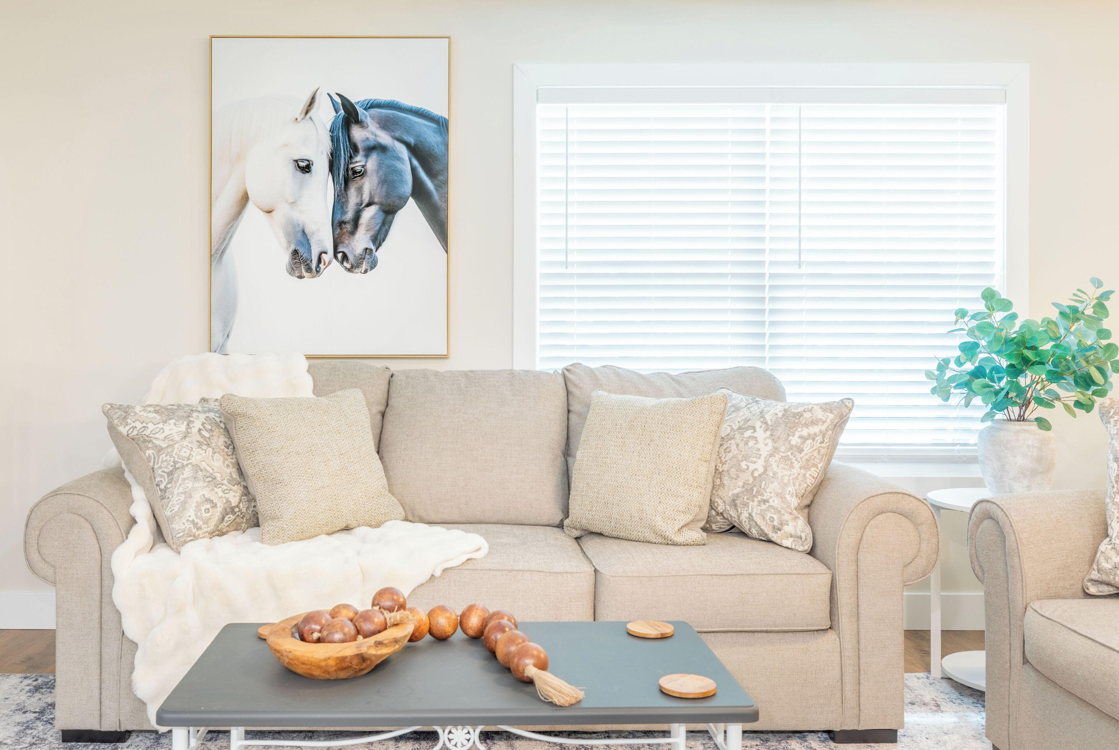 Ein Wohnzimmer mit einem beigen Sofa, einem Couchtisch mit einer Obstschale, einem Bild von einem Pferd im Rahmen und einem Fenster mit Jalousien.