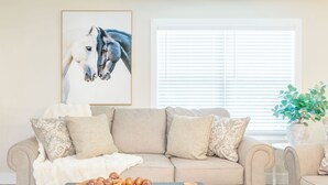 A living room with a beige sofa, a coffee table with a bowl of fruit, a framed horse painting, and a window with blinds.