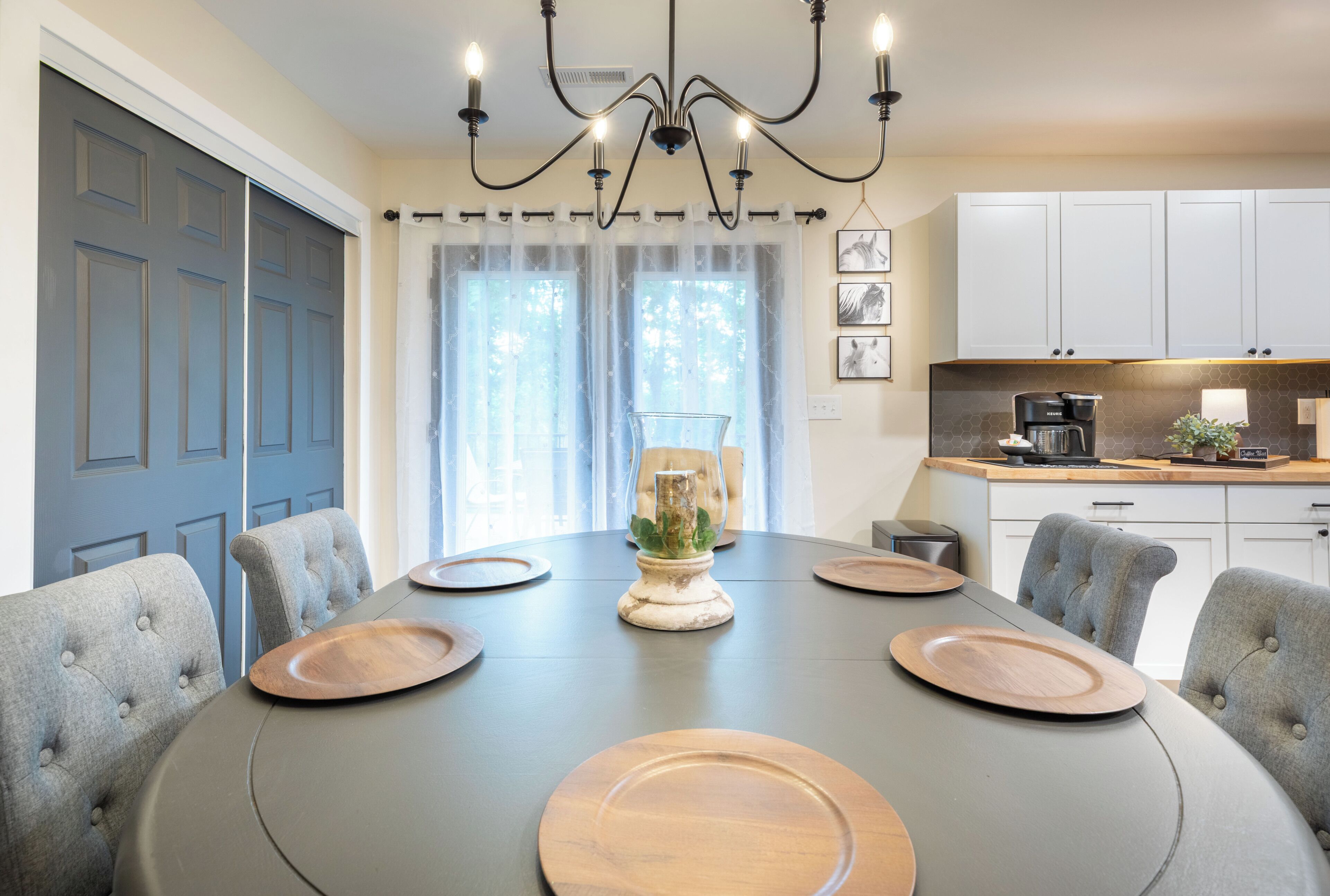 A dining area with a round table, grey upholstered chairs, a chandelier, and a kitchen in the background.