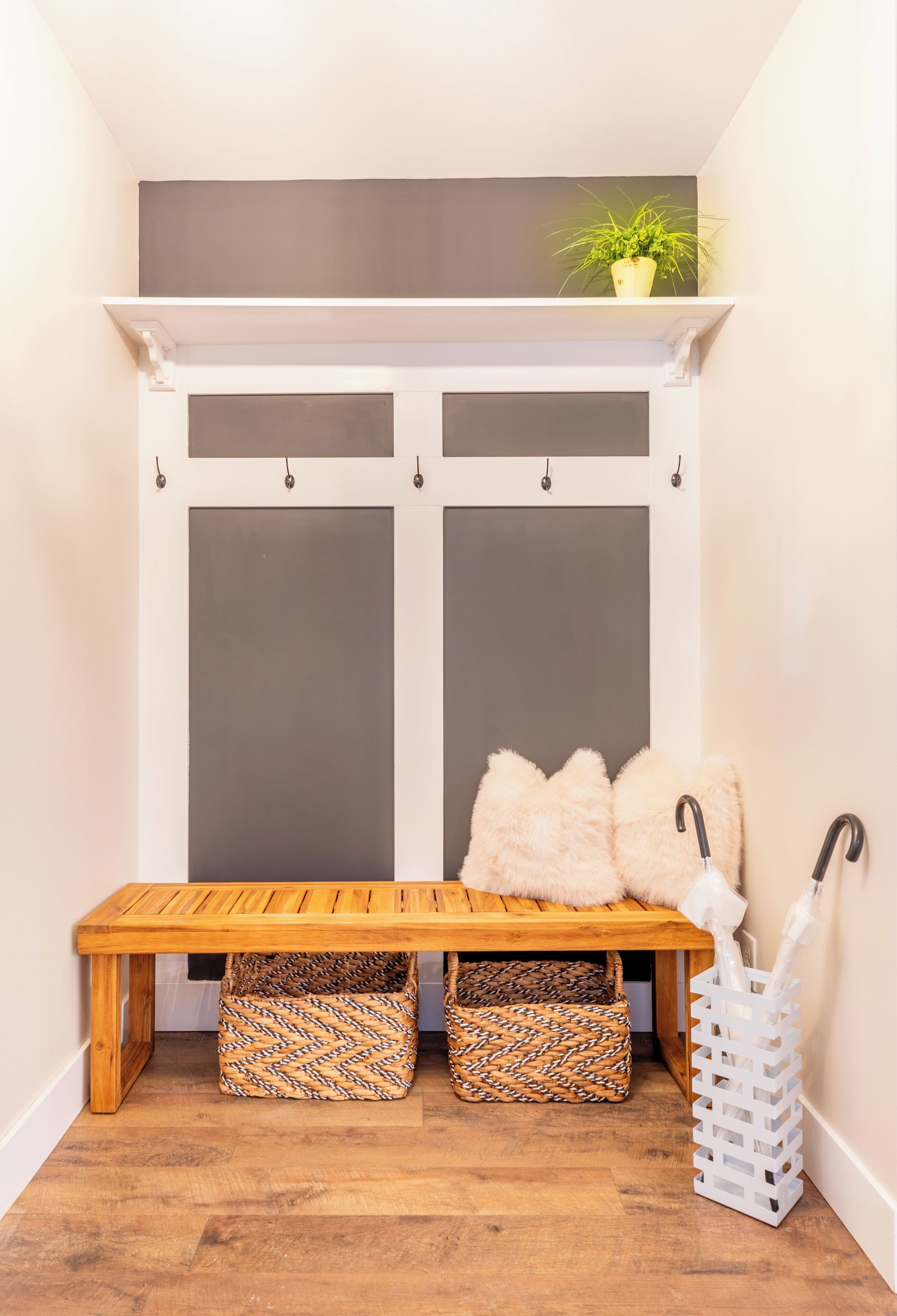 A wooden bench with storage baskets underneath, a white wall-mounted shelf with a plant, and a white basket with umbrellas.