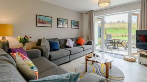 A modern living room with a grey sofa, a coffee table, and a TV. There are framed pictures on the wall, a lamp, and a view of the outdoors through a glass door. - 3 Bed in Narberth (oc-89627) (Templeton)