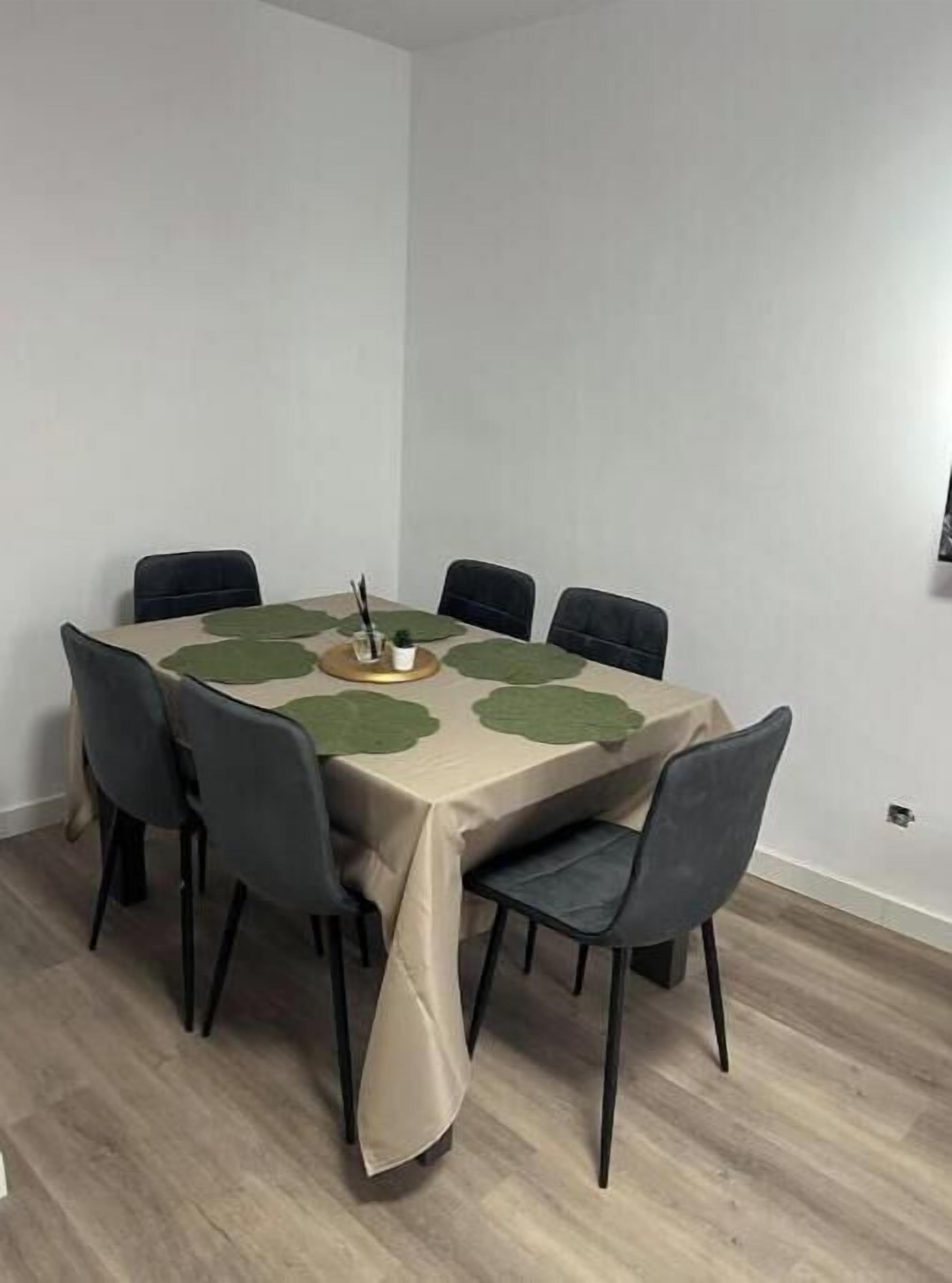 Ein rechteckiger Esstisch mit beiger Tischdecke, sechs schwarzen Stühlen und grünen Platzsets.