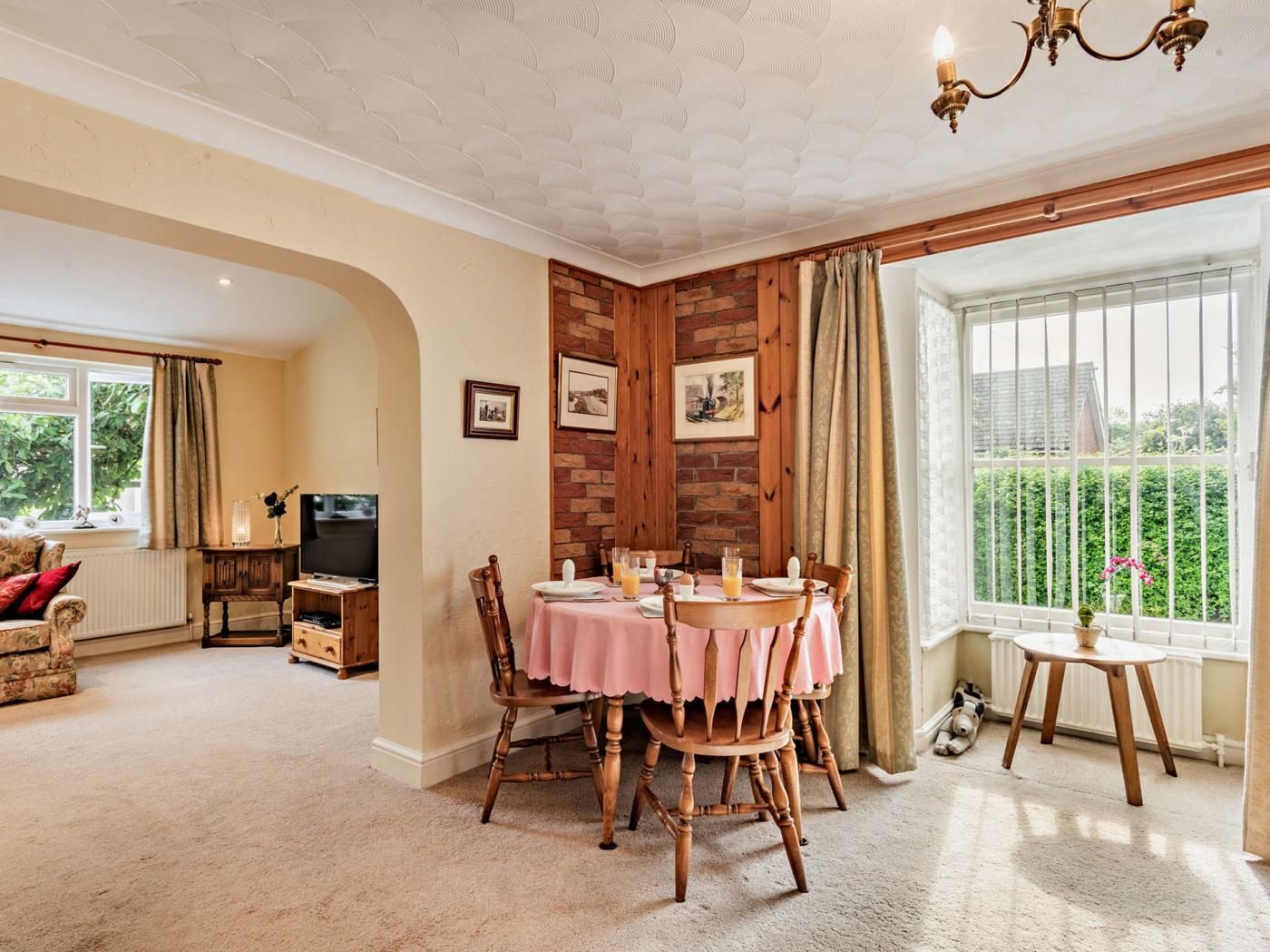 A dining area with a table set for four, wooden chairs, a chandelier, and a view of greenery through large windows.