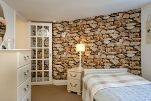 Une chambre avec un mur en pierre, un lit recouvert d’une couverture à carreaux, une table de chevet avec une lampe et une commode avec des tiroirs.
