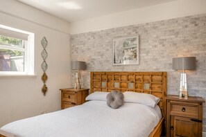 A bedroom with a wooden headboard, two bedside tables with lamps, a window, and a brick-patterned wall. - 3 Bed in Ventnor (oc-89972) (Niton)