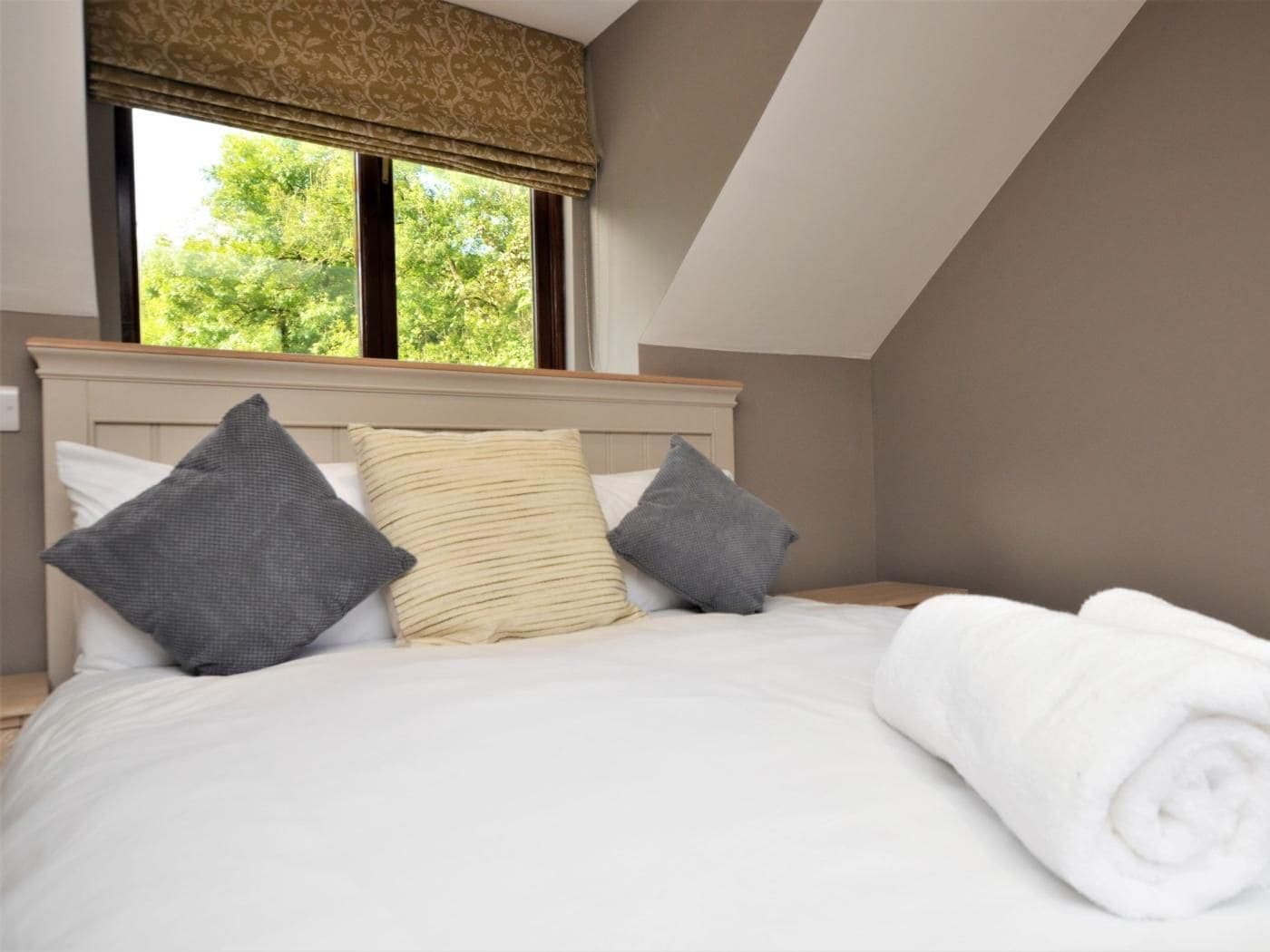 A bedroom with a bed, pillows, a window with a valance, and a view of greenery outside.