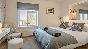 Ein Schlafzimmer mit einem Bett, einem Sessel, einem runden Hocker, einem Fenster mit Ausblick, einem Nachttisch, einer Lampe und einem Spiegel.