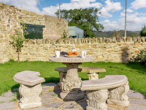 Une table en pierre, sur laquelle se trouvent une assiette de nourriture et deux tasses, est placée sur une terrasse en pierre, avec un mur de pierre en arrière-plan.
