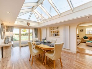 Un vaste espace à manger avec un haut plafond et une verrière, un coin salon confortable avec un canapé et des fauteuils, et une vue sur l’extérieur grâce à de grandes fenêtres.