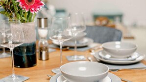Een eettafel gedekt met witte kommen, borden en bestek, met een bloevas op de achtergrond.