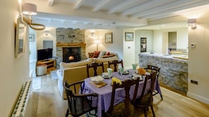 A cozy living room with a stone fireplace, a dining area with a table set for four, and a kitchen visible in the background. - 3 Bed in Leyburn (oc-89746) (Spennithorne)