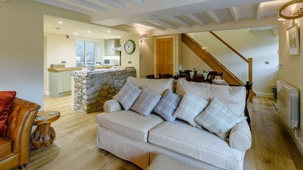 A living room with a sofa, a wooden coffee table, a stone wall, a kitchen in the background, and a staircase. - 3 Bed in Leyburn (oc-89746) (Spennithorne)