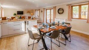 A modern kitchen with a central island, dining table with chairs, and a TV mounted on the wall. - 3 Bed in Bideford (oc-89866) (Westward Ho!)