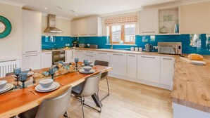 A modern kitchen with white cabinets, a wooden dining table, and blue accents. - 3 Bed in Bideford (oc-89866) (Westward Ho!)