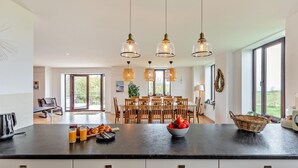 A modern kitchen with a large island, pendant lights, and a dining area with wooden chairs. - 5 Bed in Woolacombe (oc-89937) (Woolacombe)