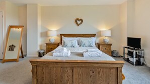 A bedroom with a wooden bed, two bedside tables with lamps, a TV stand, a mirror, and a heart-shaped wall decoration. - 5 Bed in Woolacombe (oc-89937) (Woolacombe)