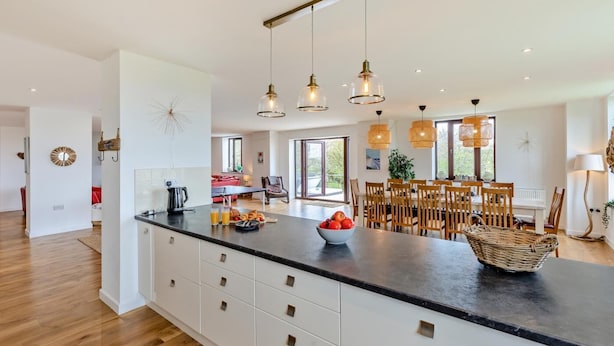 A modern kitchen with a large island, pendant lights, and a dining area with wooden chairs. - 5 Bed in Woolacombe (oc-89937) (Woolacombe)
