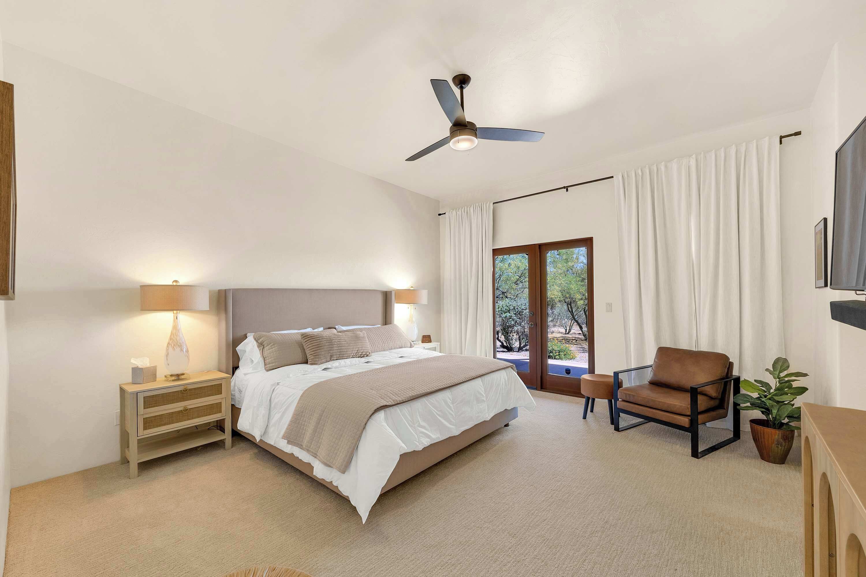 A bedroom with a bed, bedside tables, a chair, a ceiling fan, and a view through a large window.