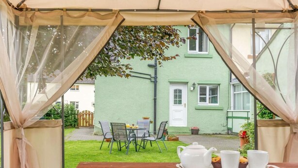 A covered outdoor seating area with a table set for tea, a green building in the background, and a tree providing shade. - 2 Bed in Hexham (oc-89299) (Kielder)